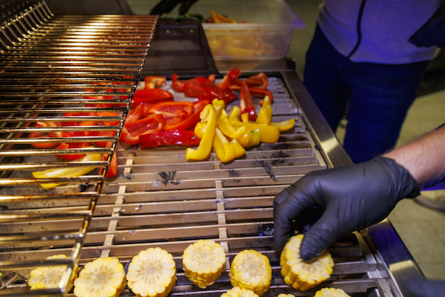Detailaufnahme von Grillgut auf einem Grillrost. Oben rote und gelbe Paprikastreifen, unten Maiskolbenstücke. Eine Hand mit schwarzem Handschuh platziert Mais auf dem Rost. Metallgrill und Teile einer Person im Hintergrund.